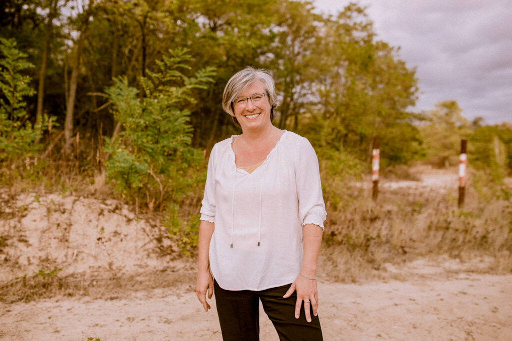 Ein Portrait der Geschäftsführerin von Strukturenwerk Martina Thum. Sie steht in lockerer Pose in der Natur und lächelt in die Kamera. Sie trägt ein bequemes weißes Oberteil und eine schwarze Hose. Ihr Haar ist mittelkurz geschnitten und Sie hat eine Brille auf.