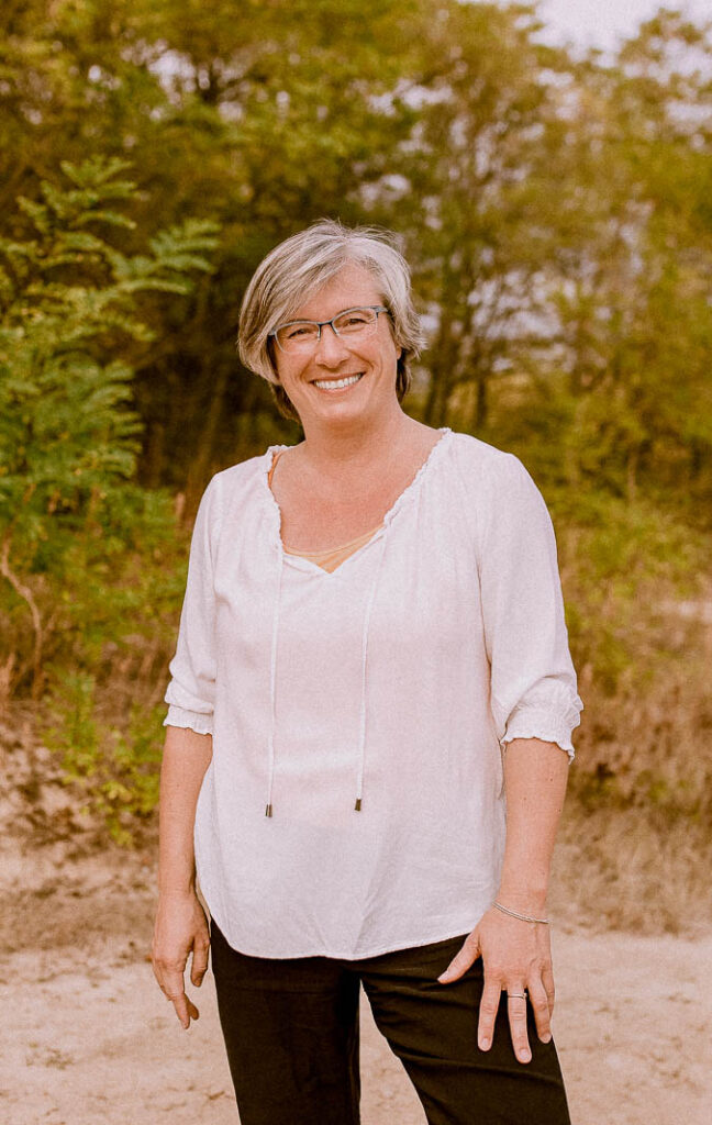 Ein Portrait der Geschäftsführerin von Strukturenwerk Martina Thum. Sie steht in lockerer Pose in der Natur und lächelt in die Kamera. Sie trägt ein bequemes weißes Oberteil und eine schwarze Hose. Ihr Haar ist mittelkurz geschnitten und Sie hat eine Brille auf.
