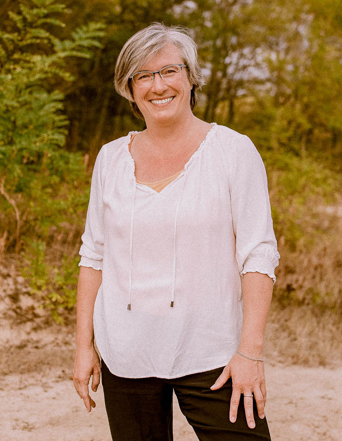 Ein Portrait der Geschäftsführerin von Strukturenwerk Martina Thum. Sie steht in lockerer Pose in der Natur und lächelt in die Kamera. Sie trägt ein bequemes weißes Oberteil und eine schwarze Hose. Ihr Haar ist mittelkurz geschnitten und Sie hat eine Brille auf.